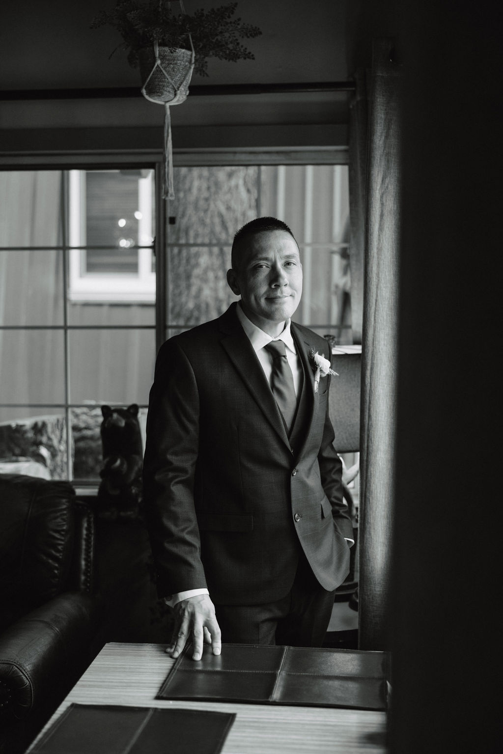 A man in a suit stands indoors by a window, with one hand resting on a table, looking intently at something out of frame. The image is in black and white.