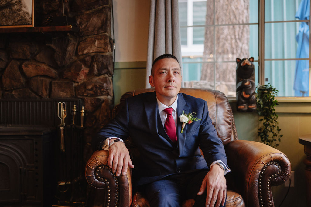 A man in a suit stands indoors by a window, with one hand resting on a table, looking intently at something out of frame. 