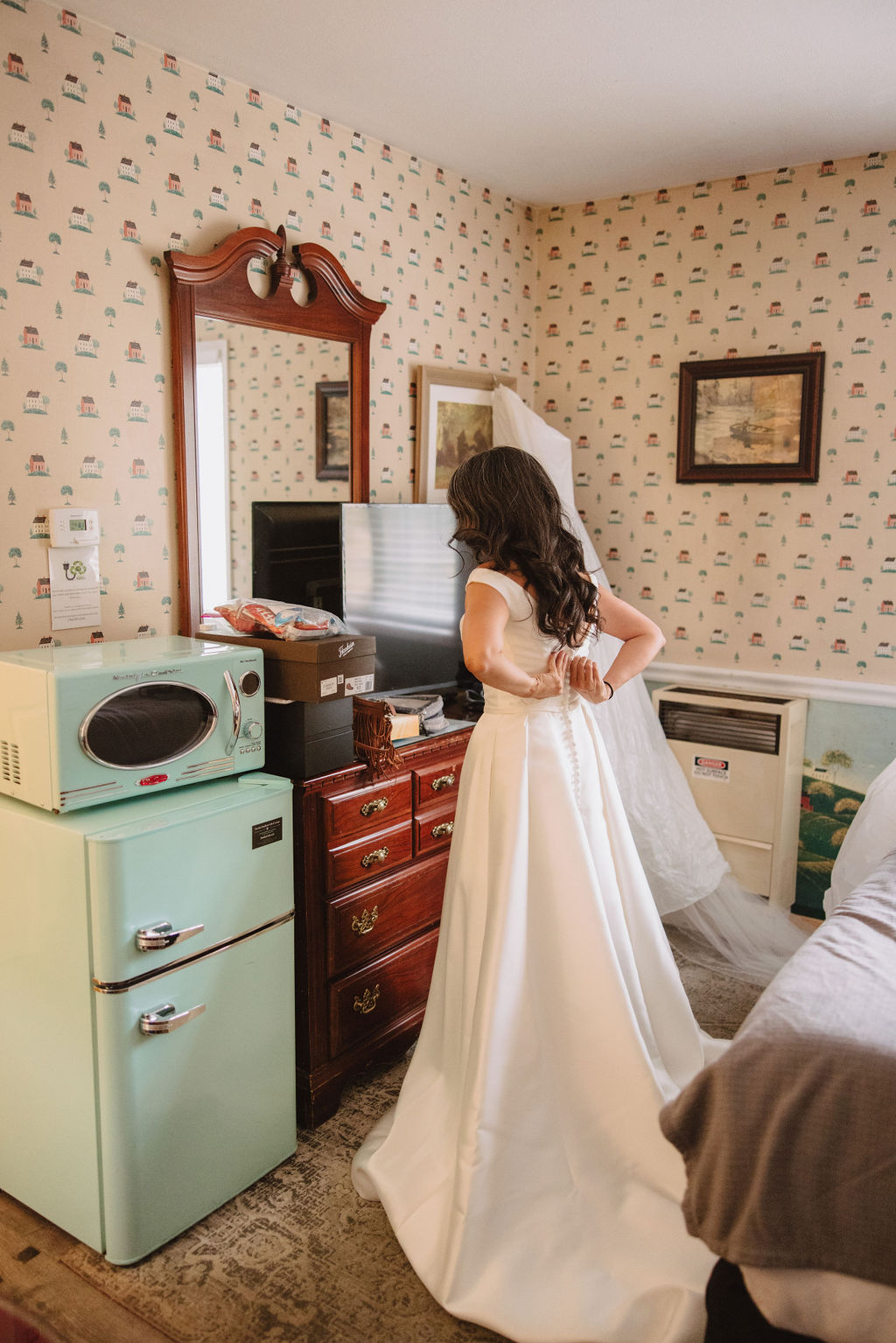 A bride, in a white gown, stands in a vintage-styled room with patterned wallpaper, adjusting her dress in front of an old wooden dresser with a mirror, a small retro fridge, and a microwave.