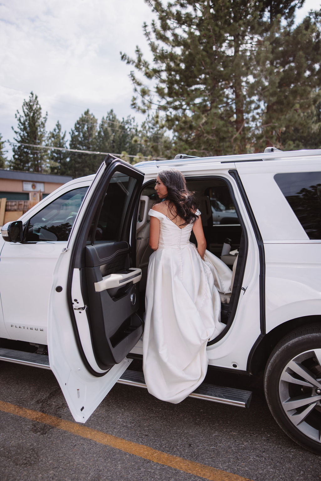 A woman in a white dress is getting into a white SUV with the rear door open in an outdoor setting. Trees and part of a building are visible in the background.