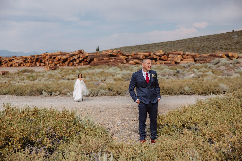 A groom in a blue suit stands in the foreground, smiling, while a bride in a white dress is seen in the background with her hands on her head in an outdoor setting for their first look at mammoth lakes 