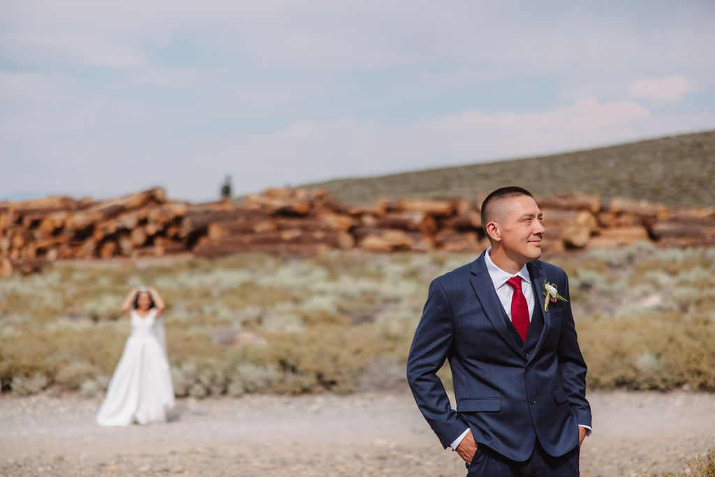 A groom in a blue suit stands in the foreground, smiling, while a bride in a white dress is seen in the background with her hands on her head in an outdoor setting for their first look at mammoth lakes 