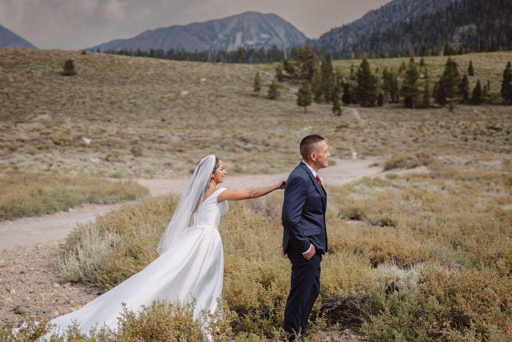 A groom in a blue suit stands in the foreground, smiling, while a bride in a white dress is seen in the background with her hands on her head in an outdoor setting for their first look at mammoth lakes 