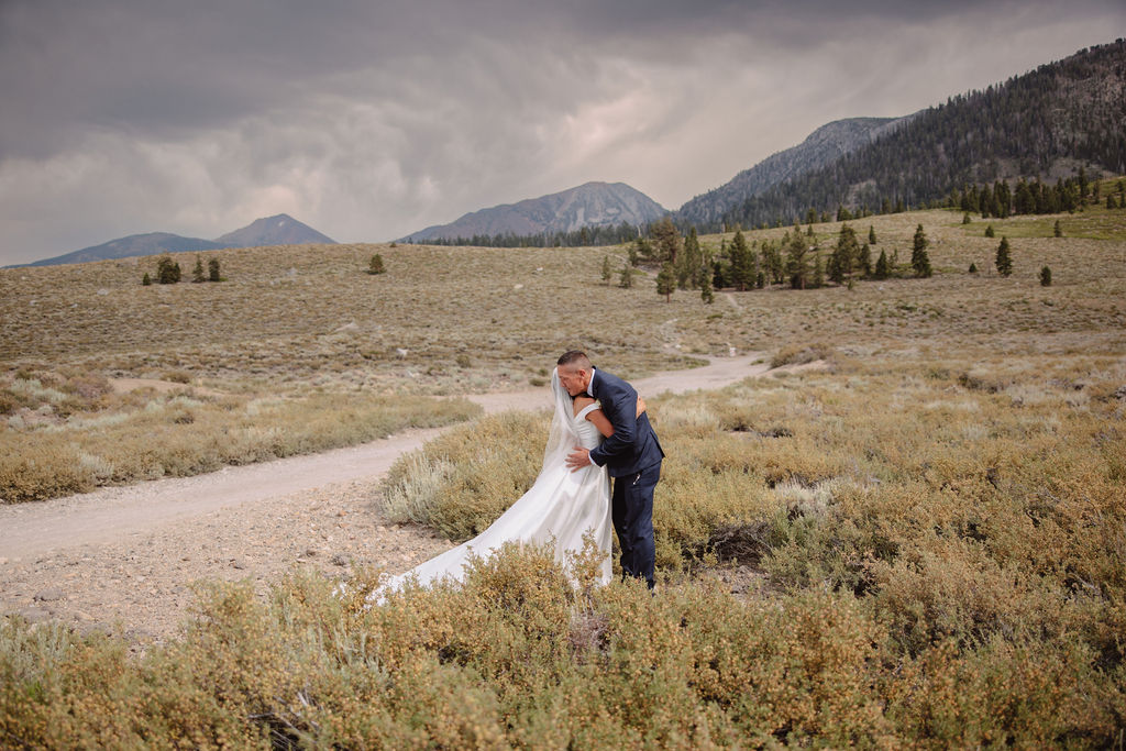 A groom in a blue suit stands in the foreground, smiling, while a bride in a white dress is seen in the background with her hands on her head in an outdoor setting for their first look at mammoth lakes 