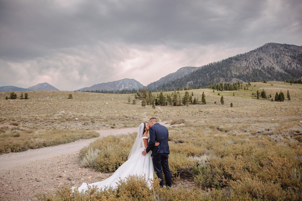 A groom in a blue suit stands in the foreground, smiling, while a bride in a white dress is seen in the background with her hands on her head in an outdoor setting for their first look at mammoth lakes