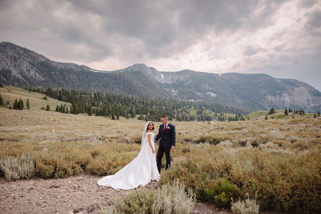 A bride in a white dress and groom in a suit stand in a vast, open field with mountains in the background, under a cloudy sky at mammoth lakes