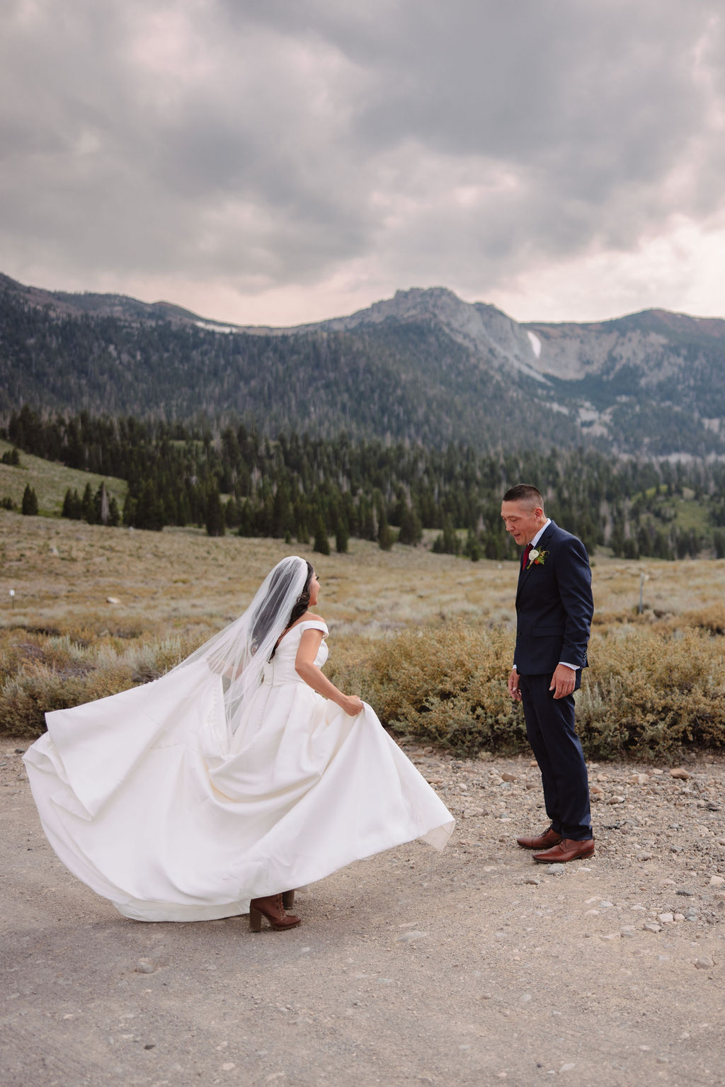 A bride in a white dress and groom in a suit stand in a vast, open field with mountains in the background, under a cloudy sky at mammoth lakes