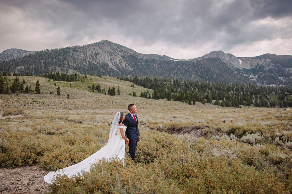 A bride in a white dress and groom in a suit stand in a vast, open field with mountains in the background, under a cloudy sky at mammoth lakes