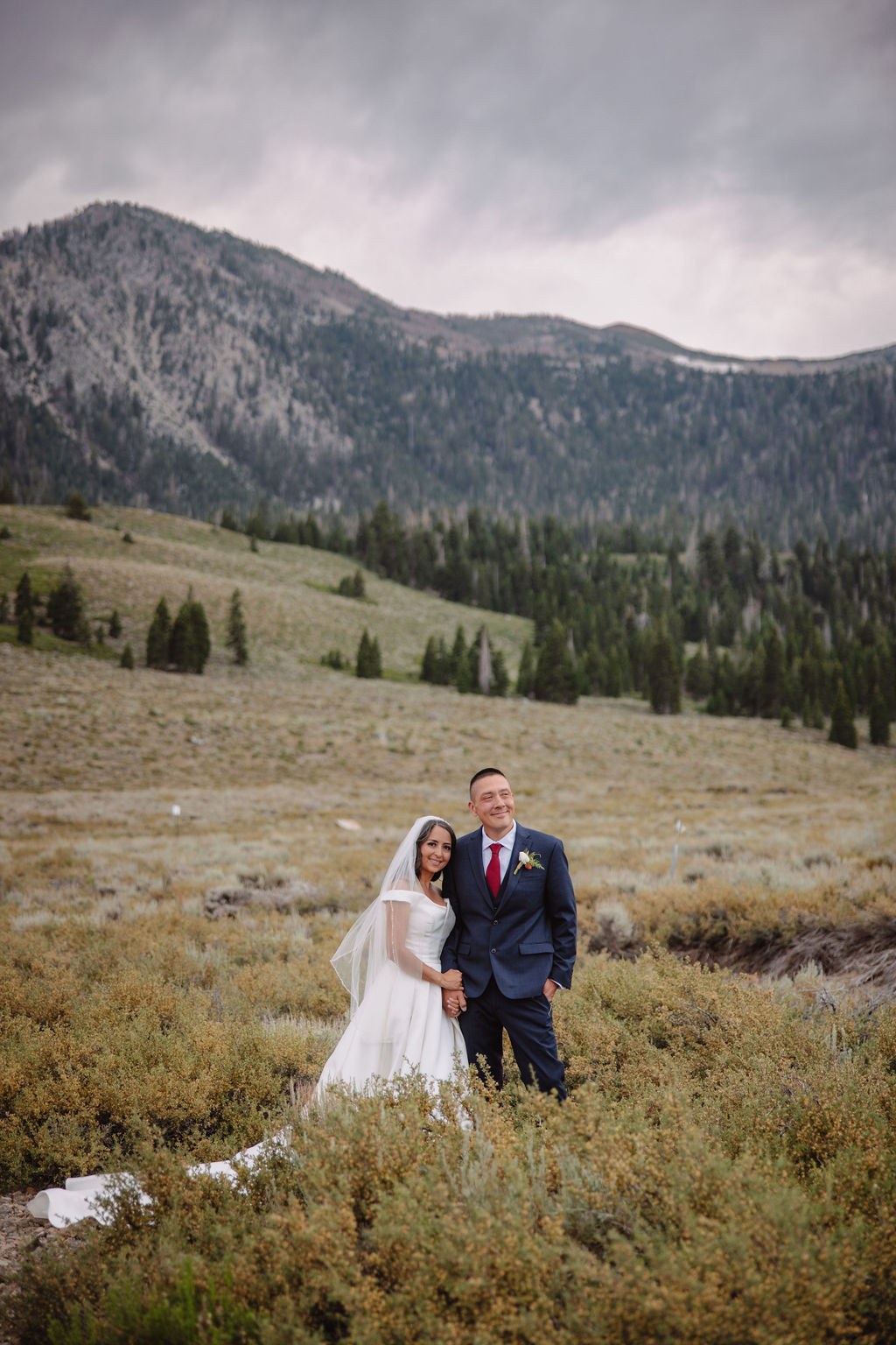 A bride in a white dress and groom in a suit stand in a vast, open field with mountains in the background, under a cloudy sky at mammoth lakes