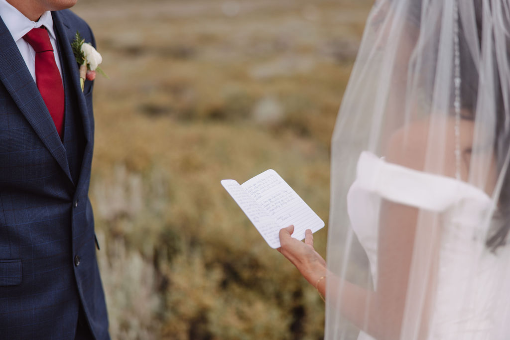 A wedding couple stands outdoors with mountains in the background. The bride, in a white gown and veil, reads from a piece of paper while the groom, in a dark suit, listens to her vows for a mammoth lakes elopement 