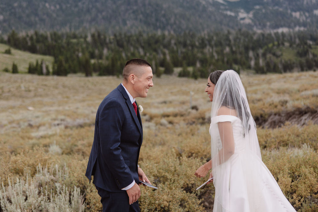 A wedding couple stands outdoors with mountains in the background. The bride, in a white gown and veil, reads from a piece of paper while the groom, in a dark suit, listens to her vows for a mammoth lakes elopement 