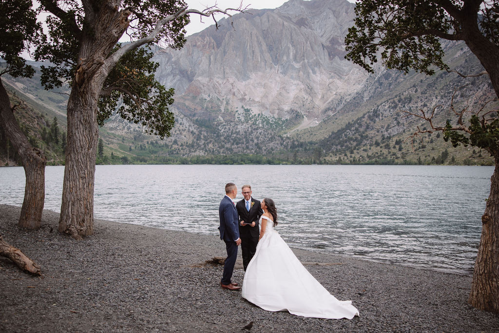 A wedding ceremony by a lakeside, with a couple in formal attire and a man officiating for a mammoth lakes elopement 