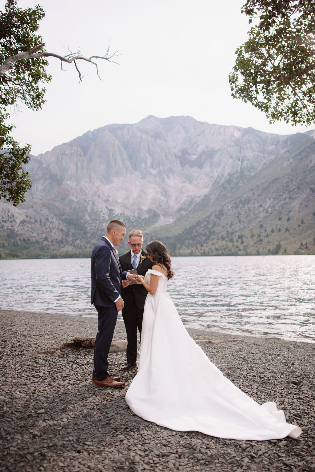 A wedding ceremony by a lakeside, with a couple in formal attire and a man officiating for a mammoth lakes elopement