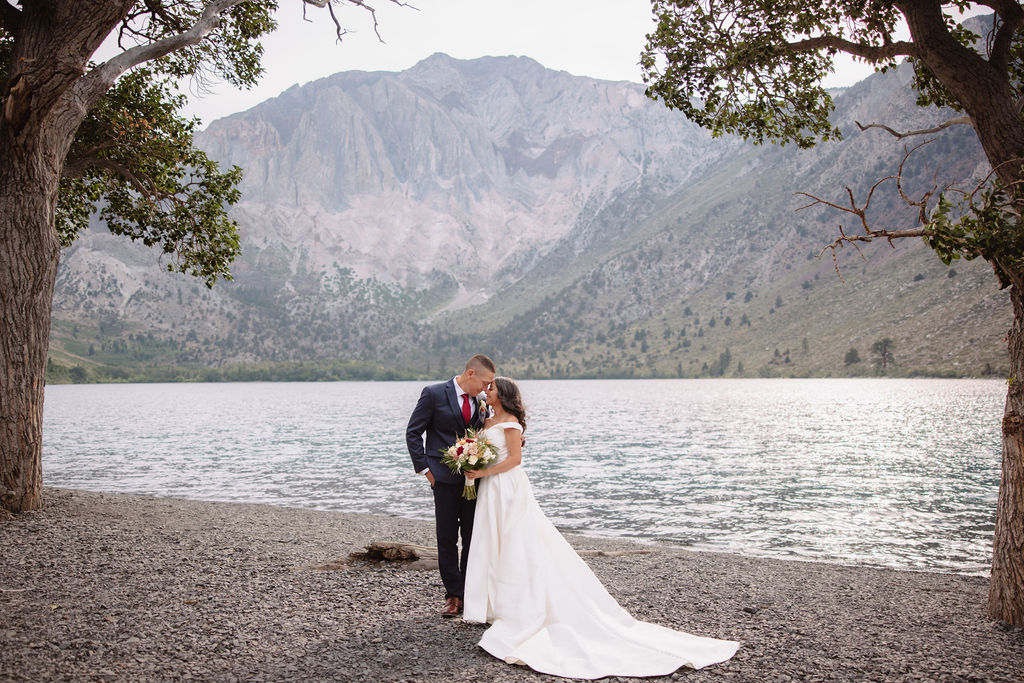 A bride and groom kiss by a lakeside, surrounded by trees and mountains in the background. The bride is holding a bouquet and is wearing a white dress. The groom is dressed in a suit.