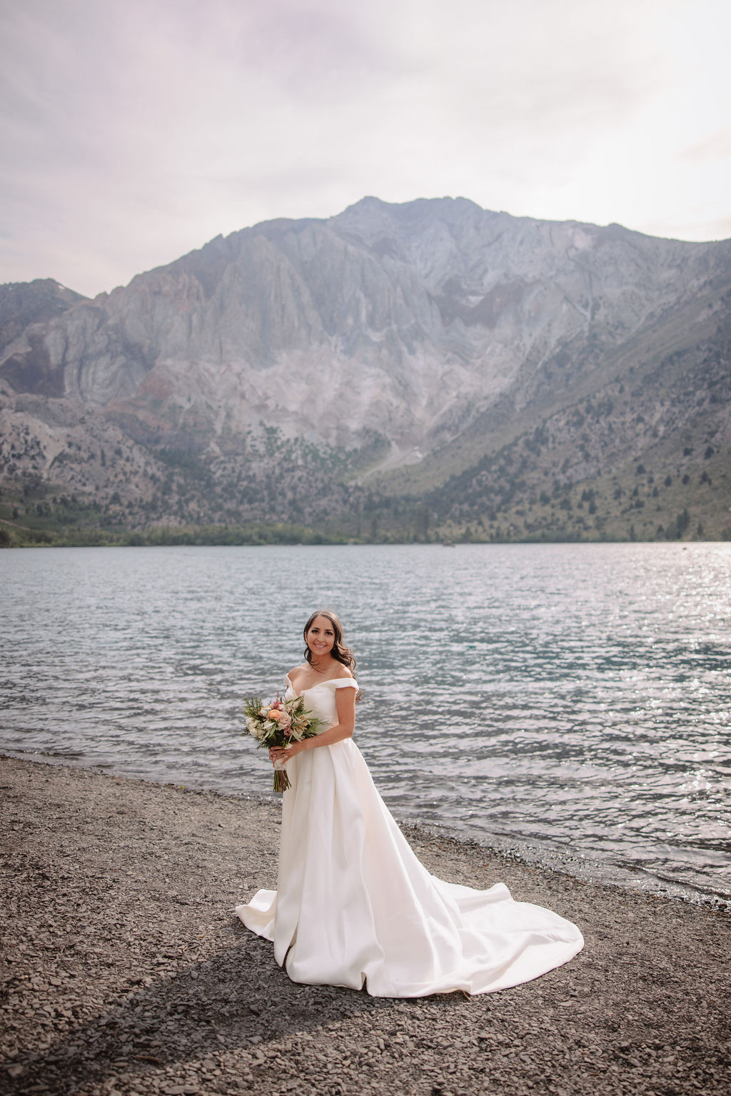 A woman in a white wedding dress holds a bouquet while standing on a rocky shore with a lake and mountains in the background.