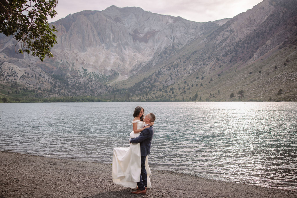 A bride in a white dress and a groom in a suit stand at the edge of a lake, with mountains in the background. He lifts her up as they gaze at each other for their mammoth lakes elopement