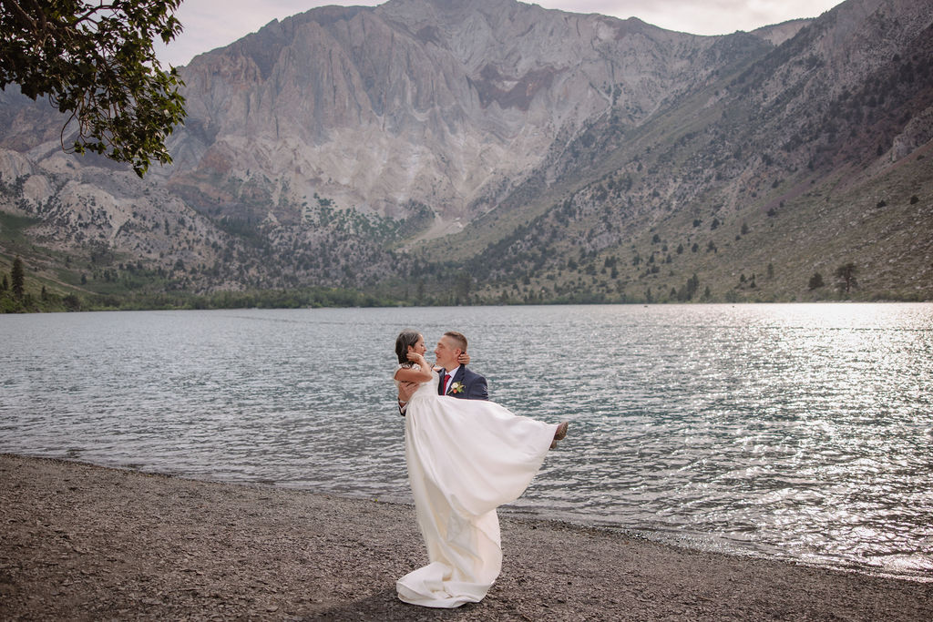 A bride in a white dress and a groom in a suit stand at the edge of a lake, with mountains in the background. He lifts her up as they gaze at each other for their mammoth lakes elopement