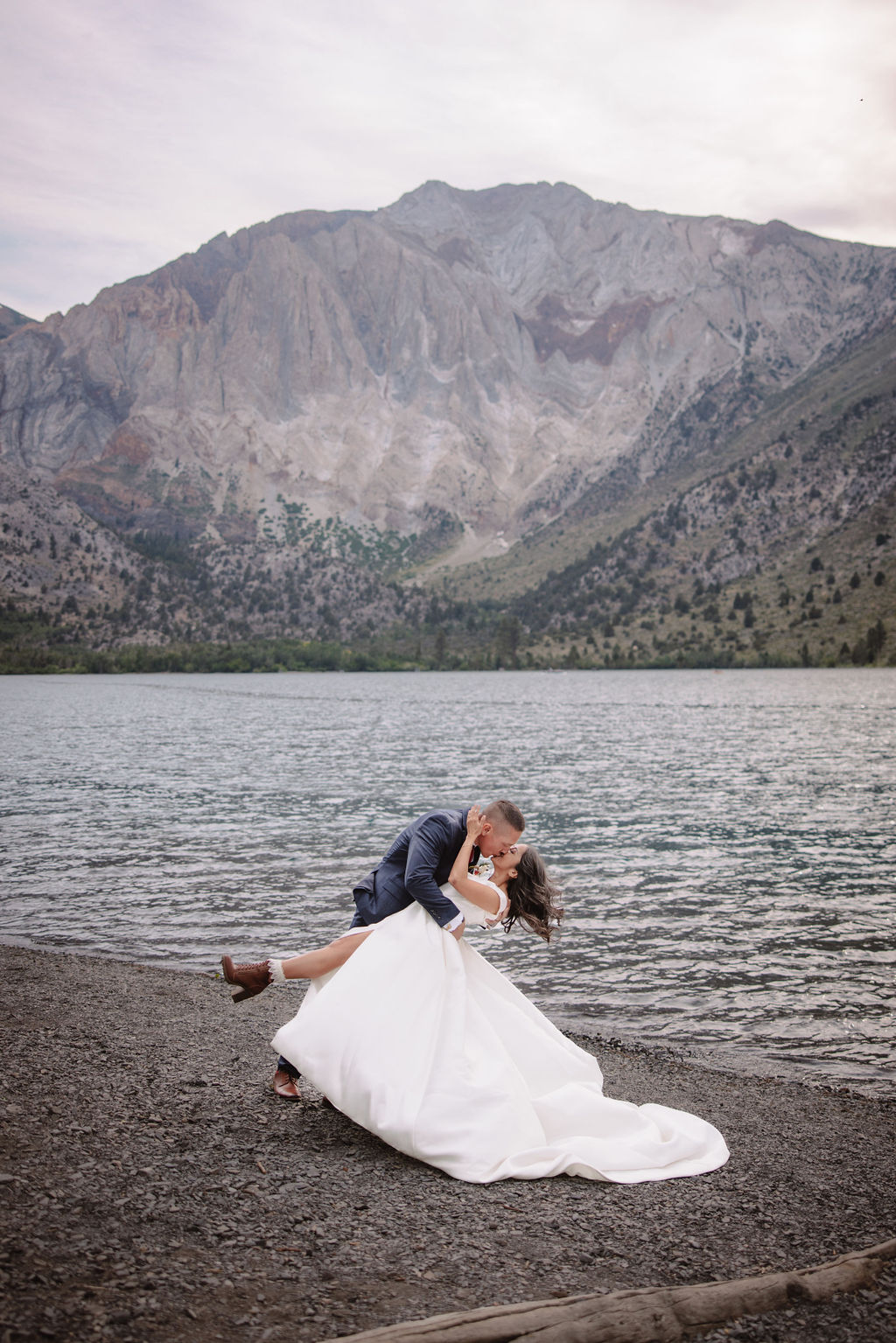 A bride in a white dress and a groom in a suit stand at the edge of a lake, with mountains in the background. He lifts her up as they gaze at each other for their mammoth lakes elopement