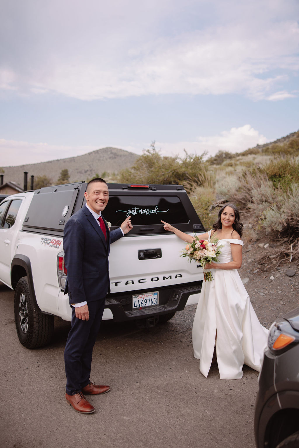 A bride and groom in wedding attire smile and point at the rear window of a white Toyota Tacoma which has "Just Married" written on it. They are outdoors with a backdrop of hills and foliage.