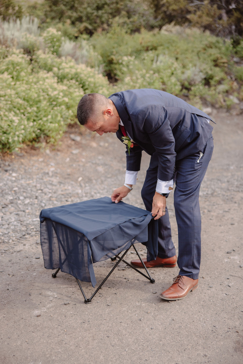 A man in a suit is setting up a small, collapsible table outdoors on a gravel path amidst greenery.