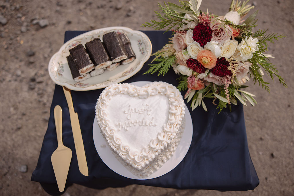 A heart-shaped white cake with the words "Just Married" inscribed on it, surrounded by piped icing. A cake server and knife are placed beside the cake on a dark tablecloth.