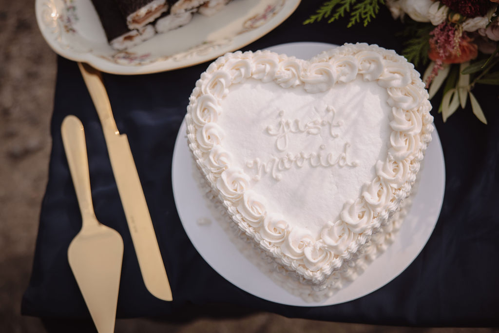 A heart-shaped white cake with the words "Just Married" inscribed on it, surrounded by piped icing. A cake server and knife are placed beside the cake on a dark tablecloth.