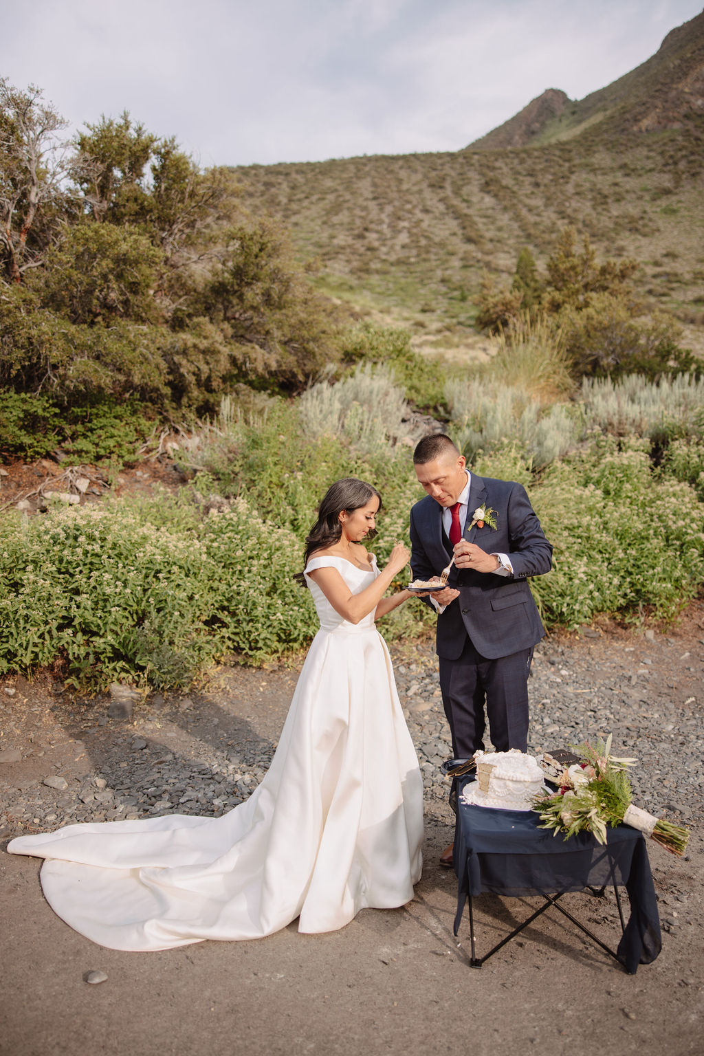 A bride in a white dress and a groom in a dark suit stand outdoors, reading from a book during their wedding ceremony, with mountains and greenery in the background.