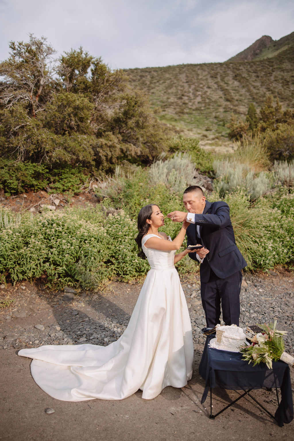 A bride in a white dress and a groom in a dark suit stand outdoors, reading from a book during their wedding ceremony, with mountains and greenery in the background.