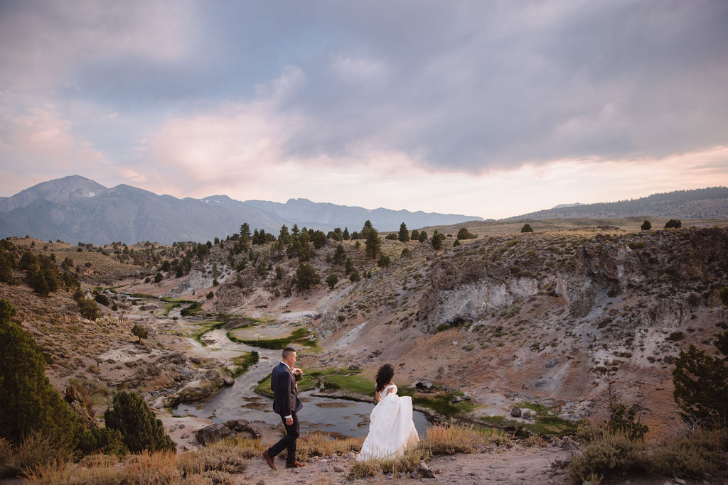 A bride and groom walk in a rocky, grassy landscape with distant mountains under a cloudy sky. The groom is standing and the bride is sitting on the ground, both looking towards the view at mammoth lakes