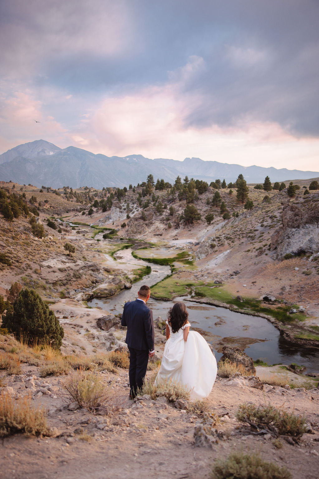 A bride and groom walk in a rocky, grassy landscape with distant mountains under a cloudy sky. The groom is standing and the bride is sitting on the ground, both looking towards the view at mammoth lakes