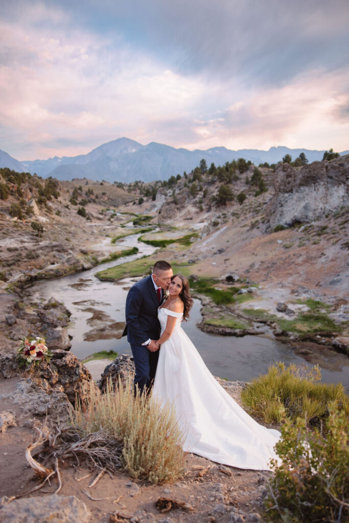 A couple standing outdoors in a rocky, mountainous landscape; the groom in a suit looks at the bride in a white dress holding a bouquet at mammoth lakes