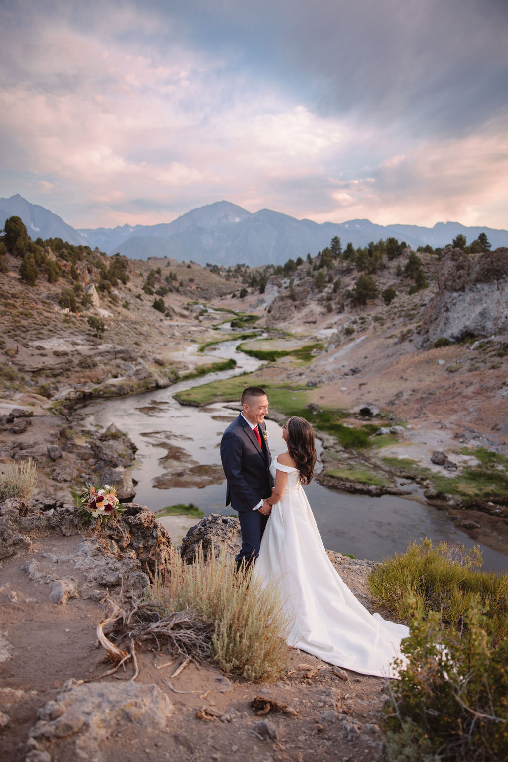 A bride and groom walk in a rocky, grassy landscape with distant mountains under a cloudy sky. The groom is standing and the bride is sitting on the ground, both looking towards the view at mammoth lakes