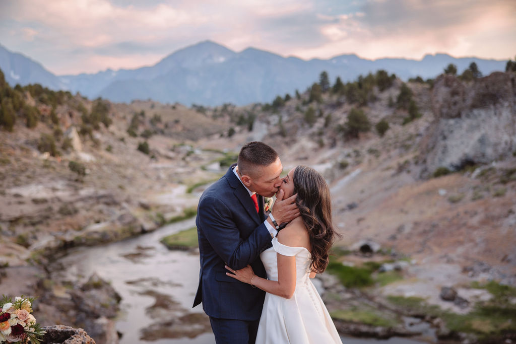 A bride and groom walk in a rocky, grassy landscape with distant mountains under a cloudy sky. The groom is standing and the bride is sitting on the ground, both looking towards the view at mammoth lakes