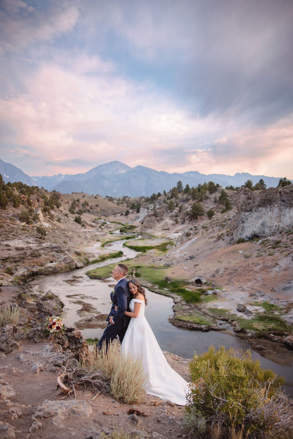 A couple standing outdoors in a rocky, mountainous landscape; the groom in a suit looks at the bride in a white dress holding a bouquet at mammoth lakes