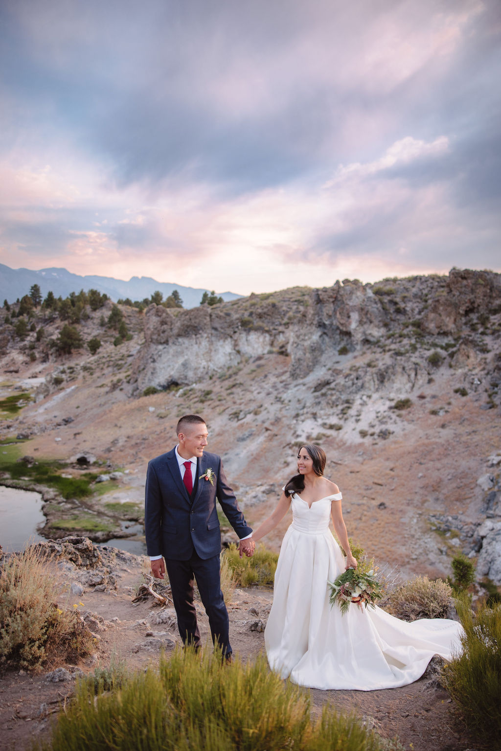 A bride and groom walk in a rocky, grassy landscape with distant mountains under a cloudy sky. The groom is standing and the bride is sitting on the ground, both looking towards the view at mammoth lakes