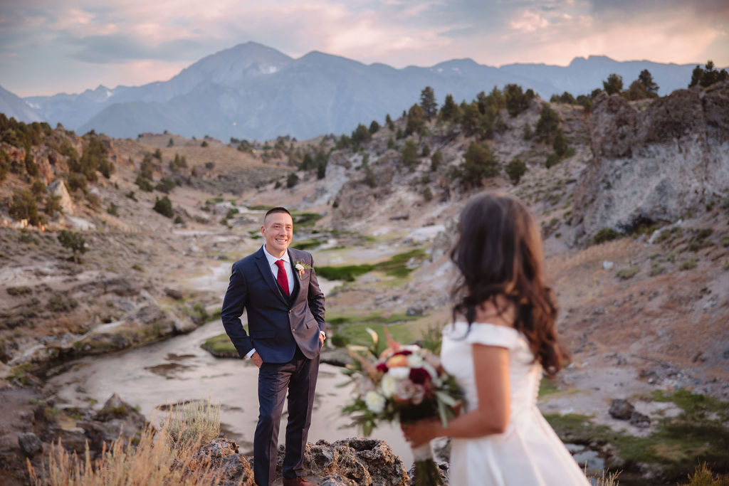 A couple standing outdoors in a rocky, mountainous landscape; the groom in a suit looks at the bride in a white dress holding a bouquet at mammoth lakes 