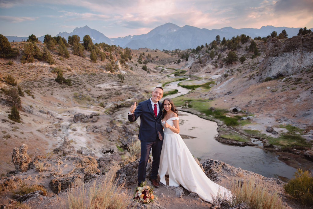 A bride and groom walk in a rocky, grassy landscape with distant mountains under a cloudy sky. The groom is standing and the bride is sitting on the ground, both looking towards the view at mammoth lakes