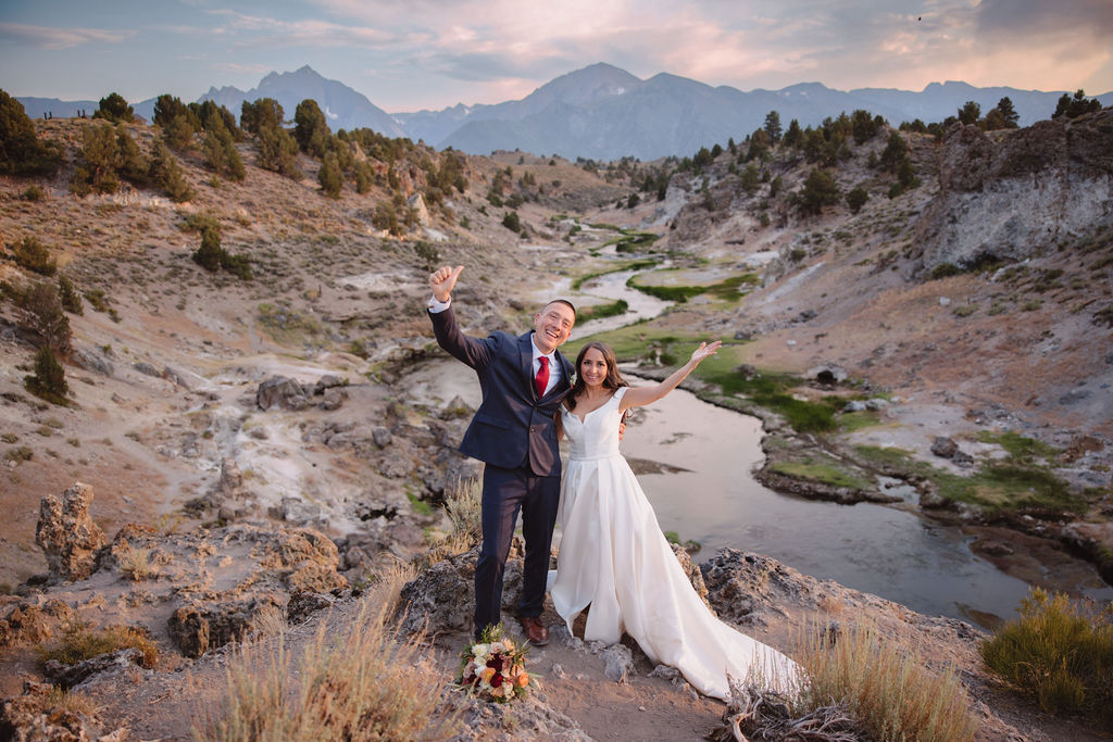 A bride and groom walk in a rocky, grassy landscape with distant mountains under a cloudy sky. The groom is standing and the bride is sitting on the ground, both looking towards the view at mammoth lakes