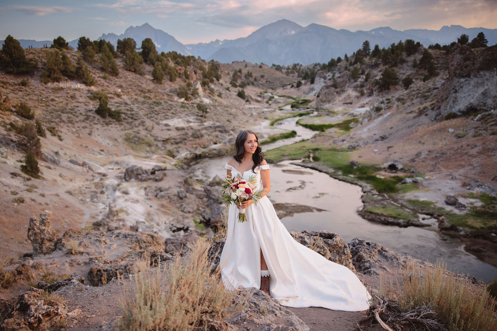 A bride and groom walk in a rocky, grassy landscape with distant mountains under a cloudy sky. The groom is standing and the bride is sitting on the ground, both looking towards the view at mammoth lakes