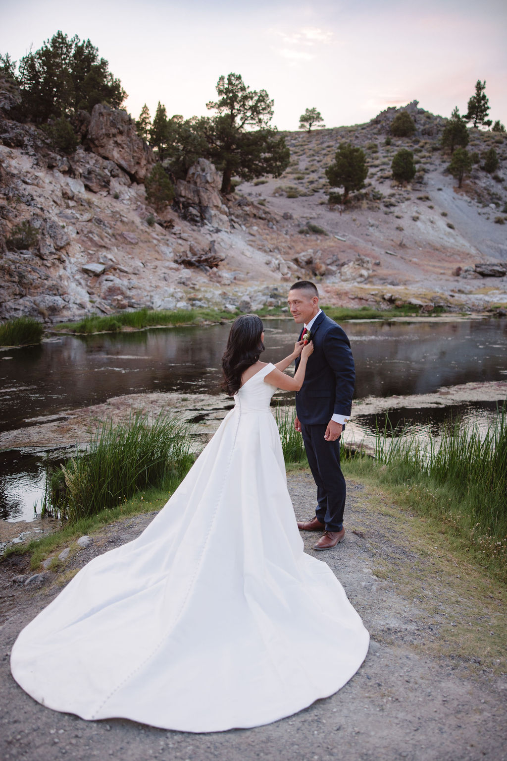 A bride and groom walk in a rocky, grassy landscape with distant mountains under a cloudy sky. The groom is standing and the bride is sitting on the ground, both looking towards the view at mammoth lakes