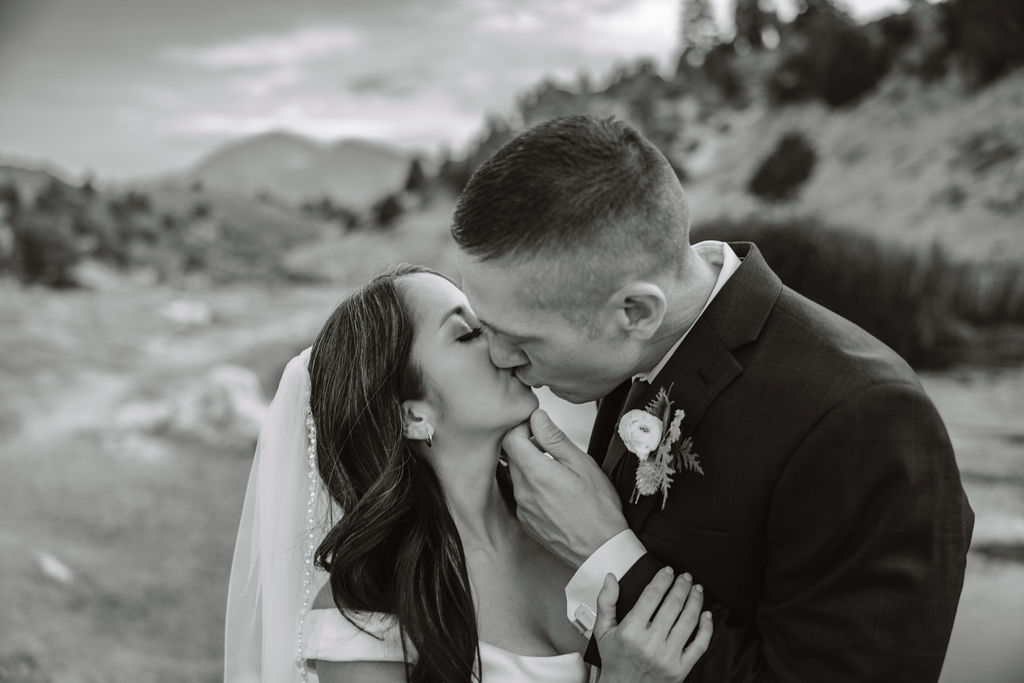 A couple standing outdoors in a rocky, mountainous landscape; the groom in a suit looks at the bride in a white dress holding a bouquet at mammoth lakes