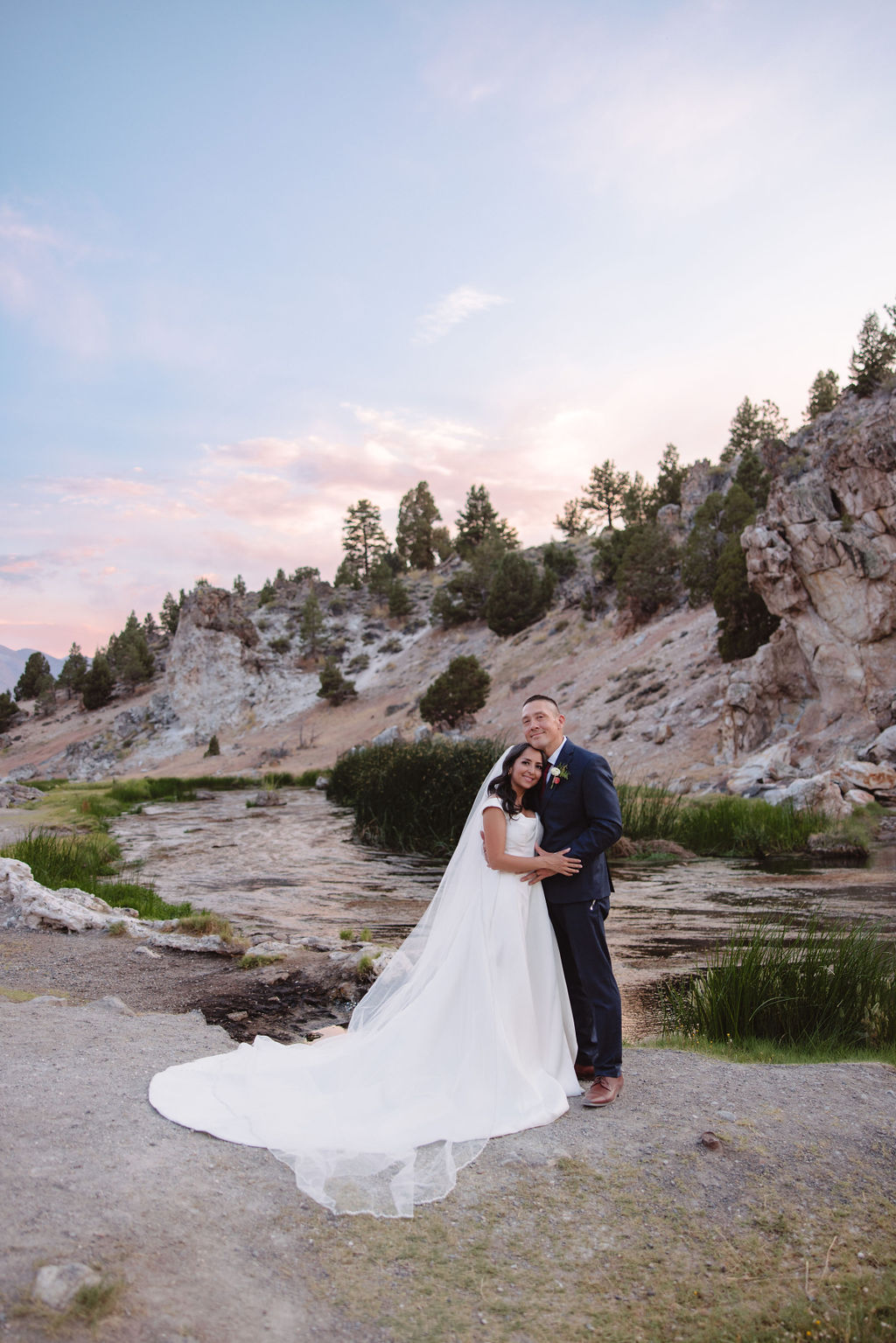 A bride and groom walk in a rocky, grassy landscape with distant mountains under a cloudy sky. The groom is standing and the bride is sitting on the ground, both looking towards the view at mammoth lakes