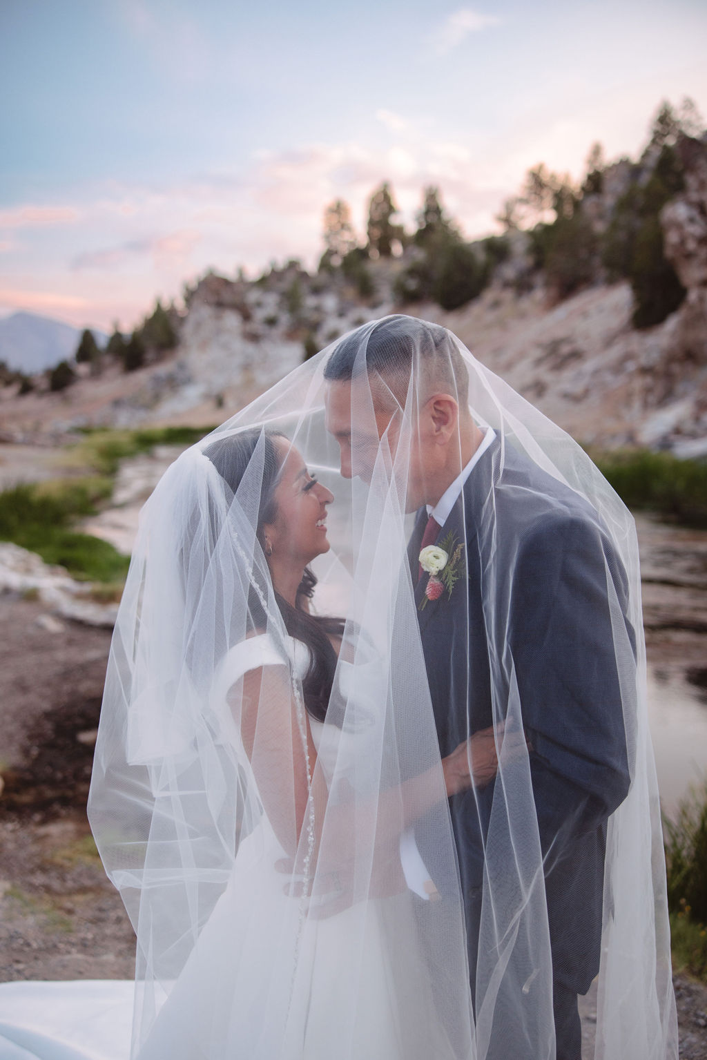 A couple standing outdoors in a rocky, mountainous landscape; the groom in a suit looks at the bride in a white dress holding a bouquet at mammoth lakes