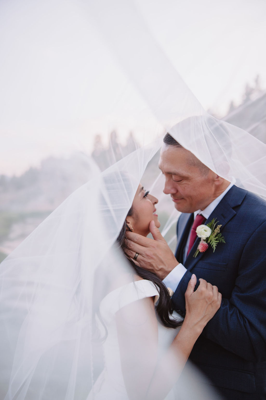 A bride and groom walk in a rocky, grassy landscape with distant mountains under a cloudy sky. The groom is standing and the bride is sitting on the ground, both looking towards the view at mammoth lakes