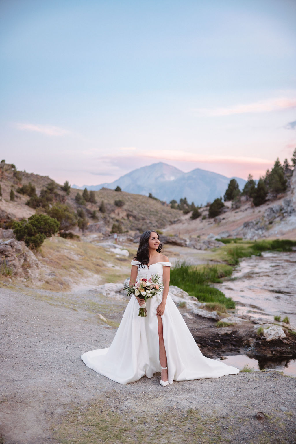 A bride and groom walk in a rocky, grassy landscape with distant mountains under a cloudy sky. The groom is standing and the bride is sitting on the ground, both looking towards the view at mammoth lakes