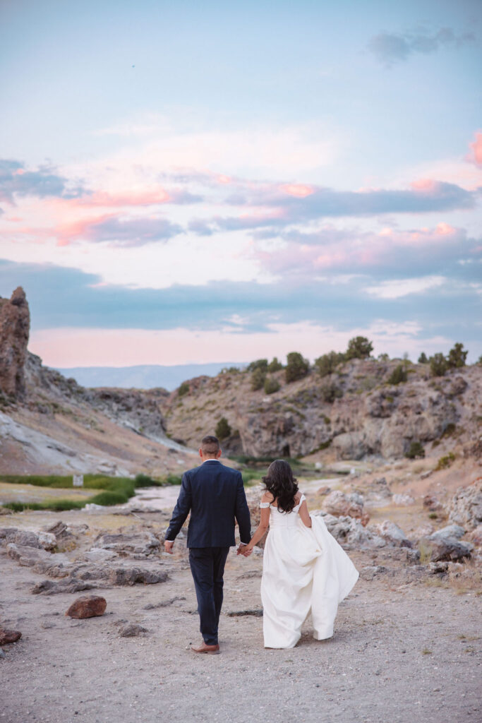 A bride and groom walk in a rocky, grassy landscape with distant mountains under a cloudy sky. The groom is standing and the bride is sitting on the ground, both looking towards the view at mammoth lakes