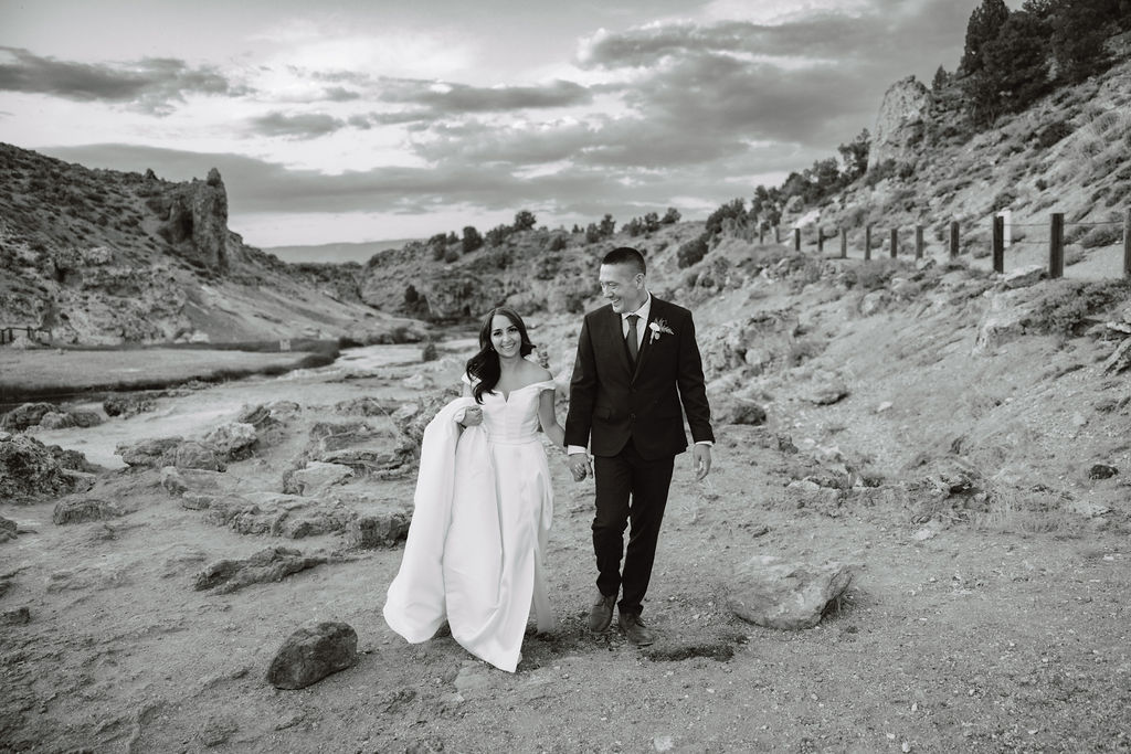 A bride and groom walk in a rocky, grassy landscape with distant mountains under a cloudy sky. The groom is standing and the bride is sitting on the ground, both looking towards the view at mammoth lakes