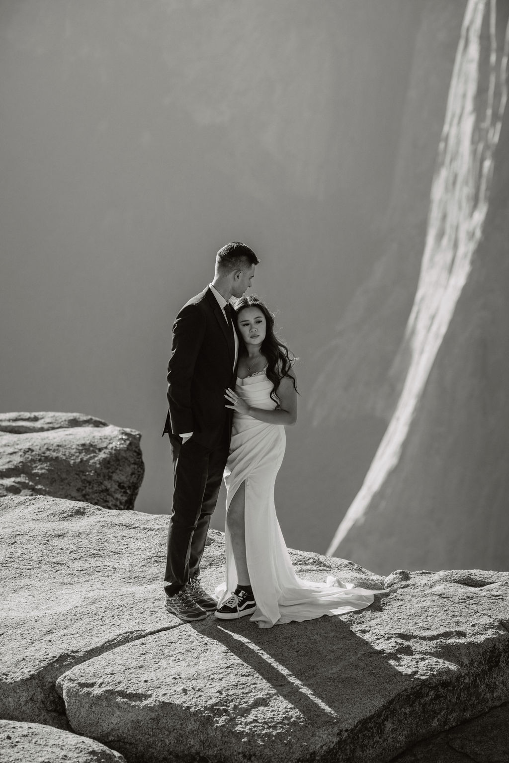 A couple in formal attire stands on a rocky outcrop with a waterfall in the background. The man wears a suit and the woman is in a strapless gown. It appears to be a wedding photo.