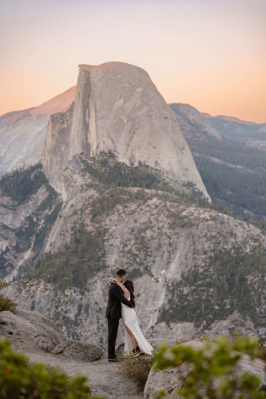 A couple in formal attire embraces on a rocky ledge with a large mountain peak in the background at sunset.