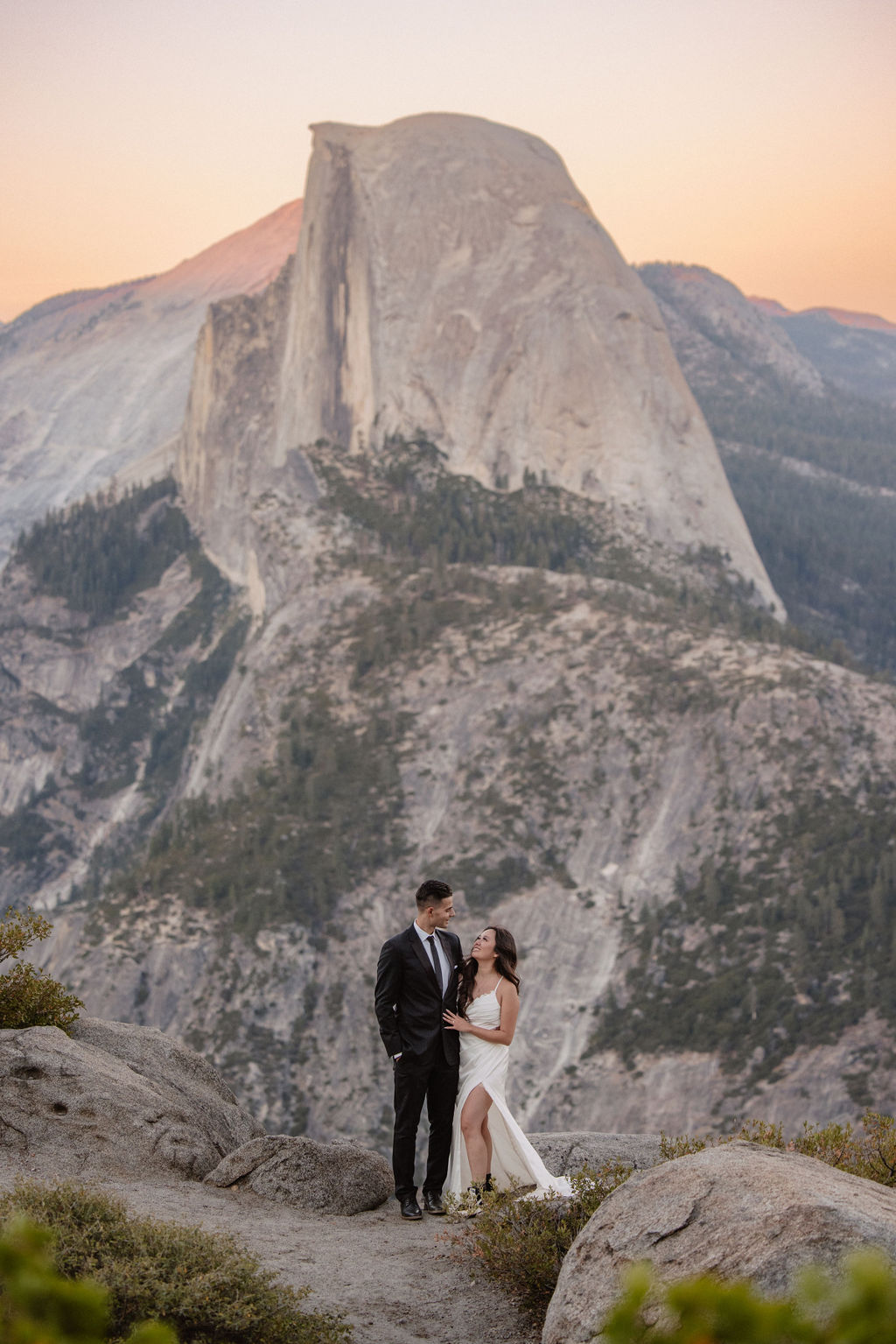 A couple in formal attire embraces on a rocky ledge with a large mountain peak in the background at sunset.